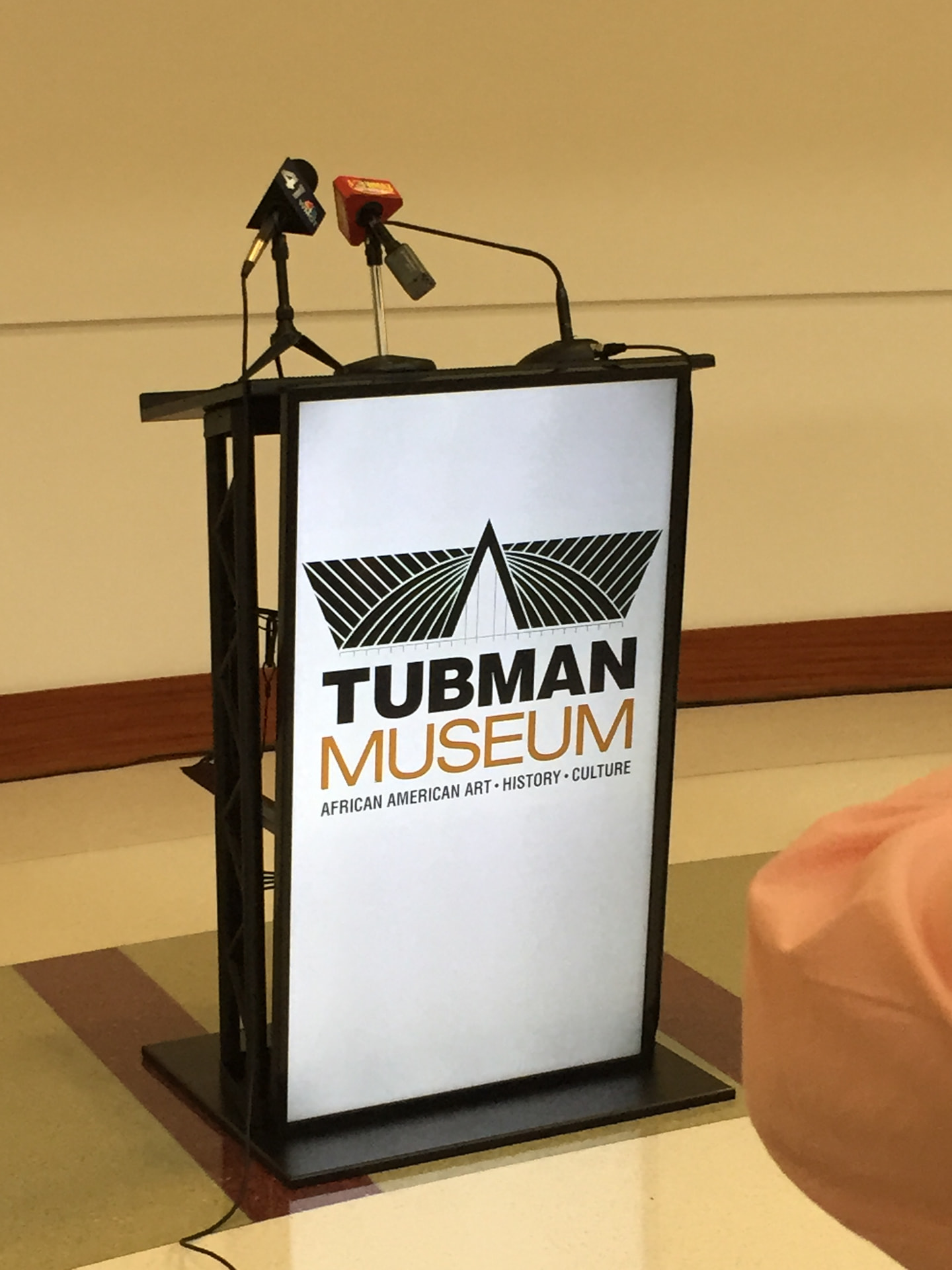 Lit truss podium with branded panel and press microphones at a museum press conference